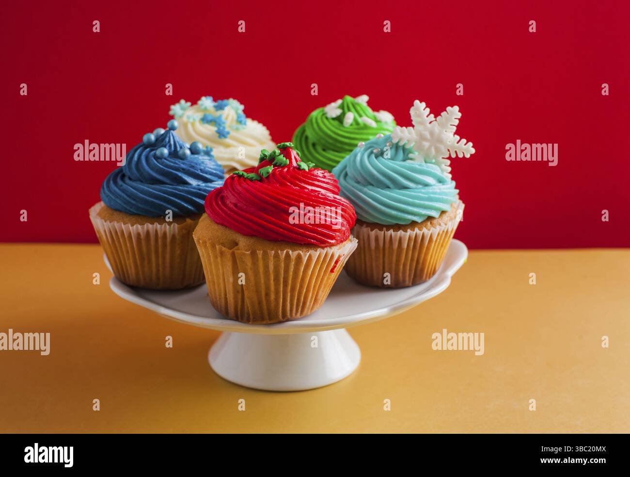 Weißer Kakestand mit fünf Cupcakes mit Sahne bedeckt, auf einem Holztisch. Jeder Cupcake hat seine eigene farbige Glasur oben drauf. Isoliert auf rotem Hintergrund Stockfoto