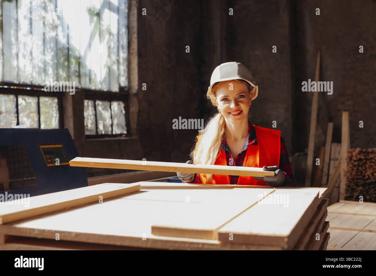 Porträt einer Holzarbeiterin mit Holzplanke. Lächelnde Arbeiterin Uniform und Helm, lächelnd, in die Kamera schauend Stockfoto