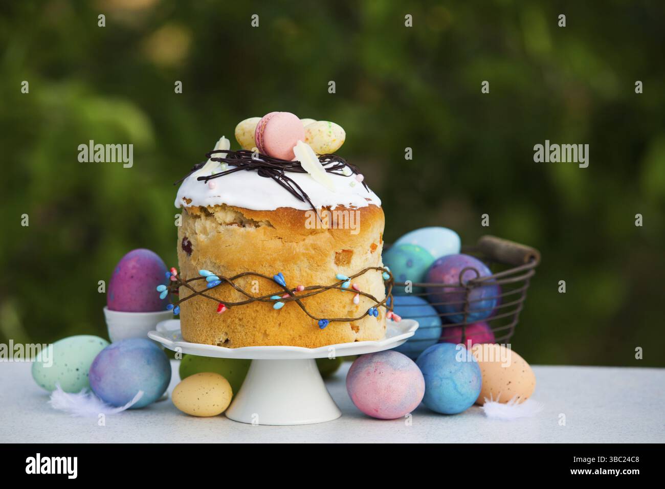 Foto von leckerem Osterbrot mit originaler Dekoration aus weißem Glasur, rosafarbenen Makronen und Schokoladenstroh. Große Menge an stilvollen, farbigen Eiern in der Nähe von cak Stockfoto