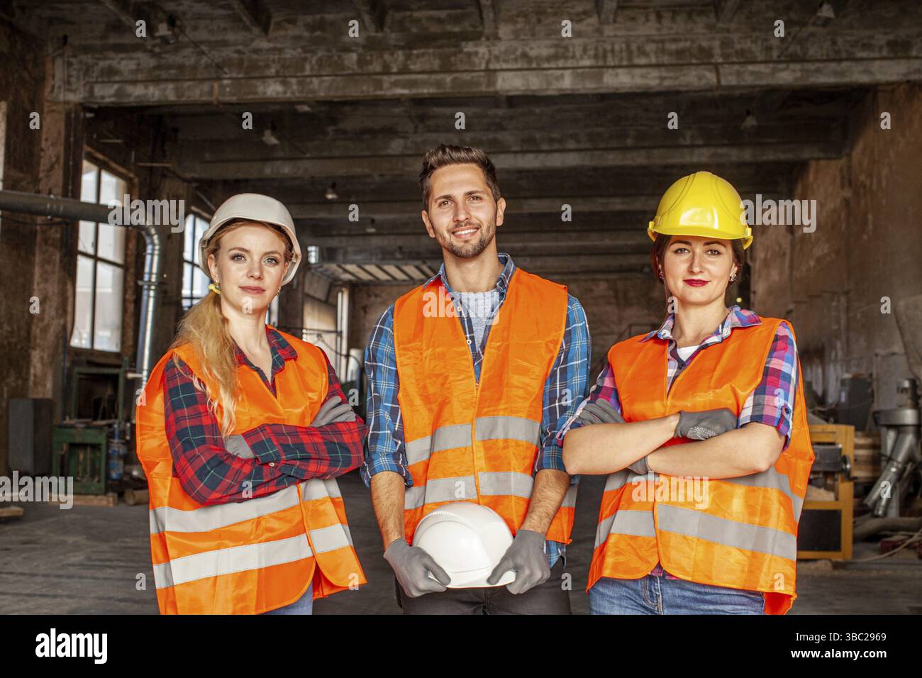 Zwei Frauen und ein Mann, junge Fabrikarbeiter in orangefarbener Schutzweste und Schutzhelmen bei der Arbeit in alten Fabriken. Frauen stehen mit gekreuzter Hand Stockfoto