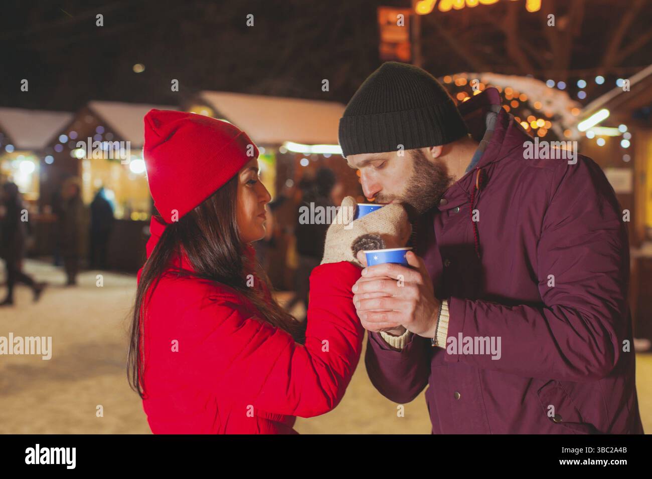 Attraktive junge Liebhaber am Abend Weihnachtsmarkt. Schöner mann Holding heiße Tasse mit beiden Händen, schöne Frau in Rot Winterkleidung behandelt ihre spous Stockfoto