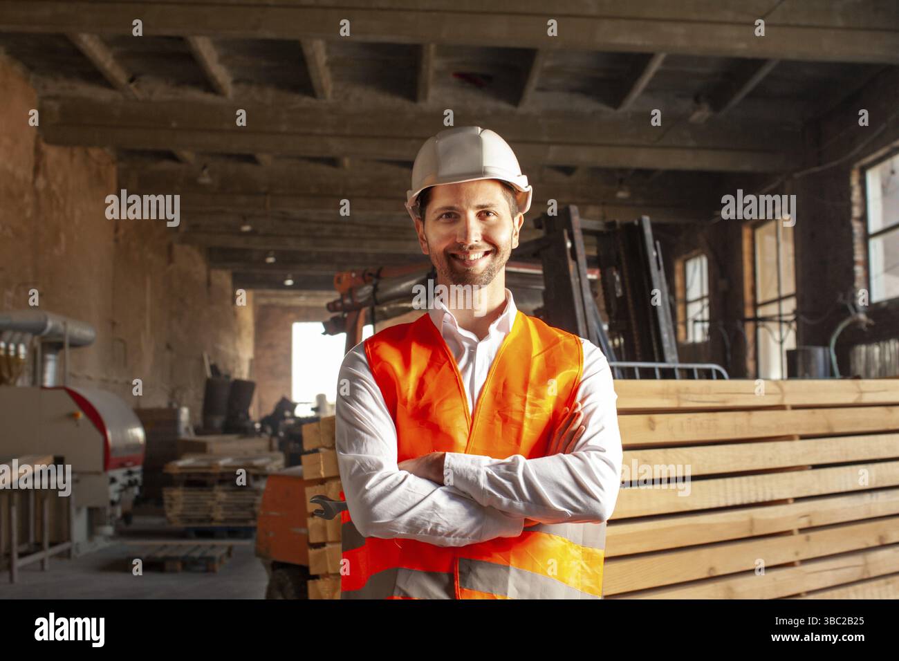 Junger lächelnder und glücklicher Ingenieur in orangefarbener Weste und Schutzhelm. Holzarbeiter im Werk Stockfoto