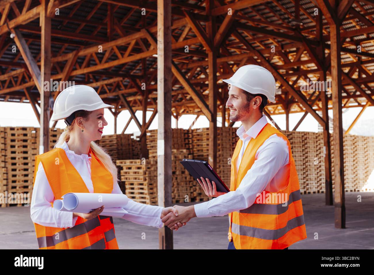 Eine attraktive junge Frau, die sich die Hand schüttelt, während ein professioneller Baumeister im Freien auf dem Hintergrund der Holzpalettenlagerung steht. Letzte Phase der Vereinbarung Stockfoto