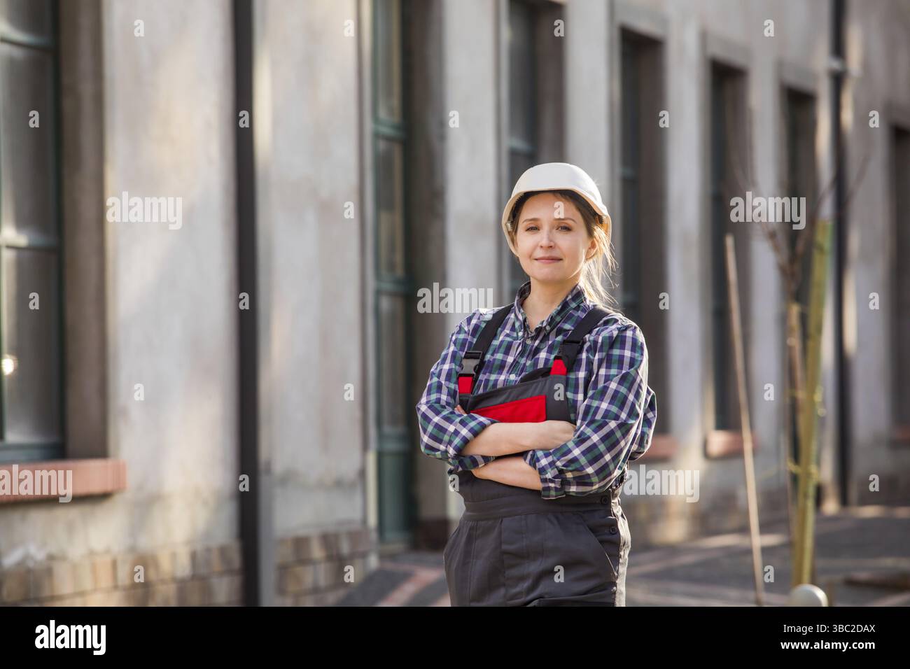 Porträt einer glücklichen, selbstbewussten Arbeiterin mit weißem Schutzhelm und Arbeitskleidung, Posen mit gefalteten Händen auf der Brust. Junge Profis sind stolz Stockfoto