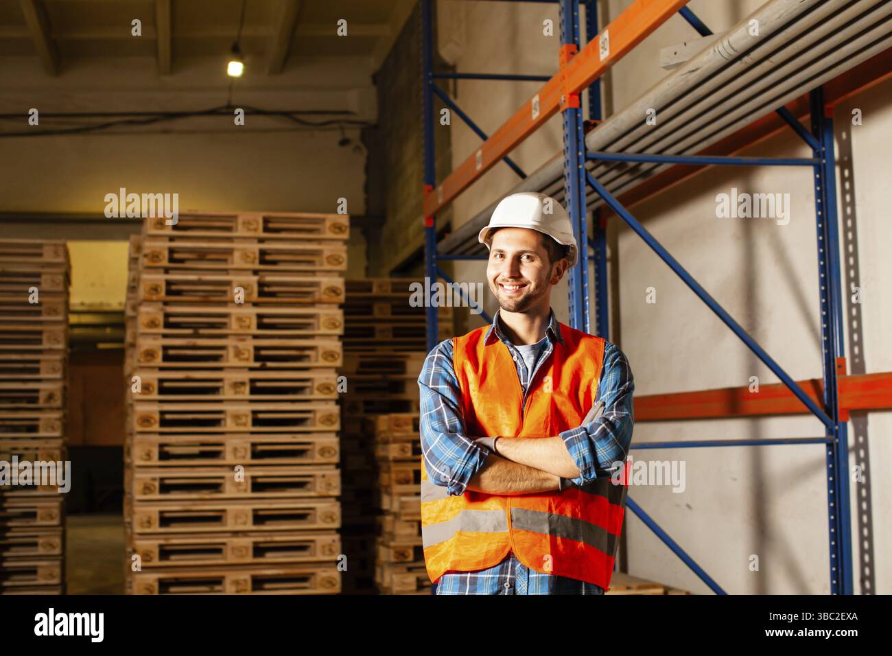 Glücklicher junger Arbeiter in Schutzuniform vor Holzpaletten. Palettenstapel im Lager Stockfoto
