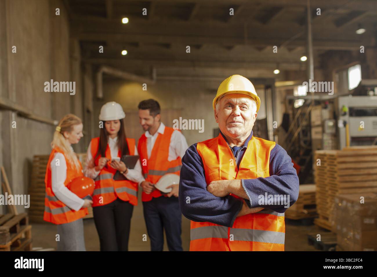 Team junger Geschäftsleute in orangefarbenen Schutzwesten und Schutzhelmen, die in der Gruppe auf dem Hintergrund stehen, reifer Vorarbeiter, der Hände an der Brust hält, St. Stockfoto