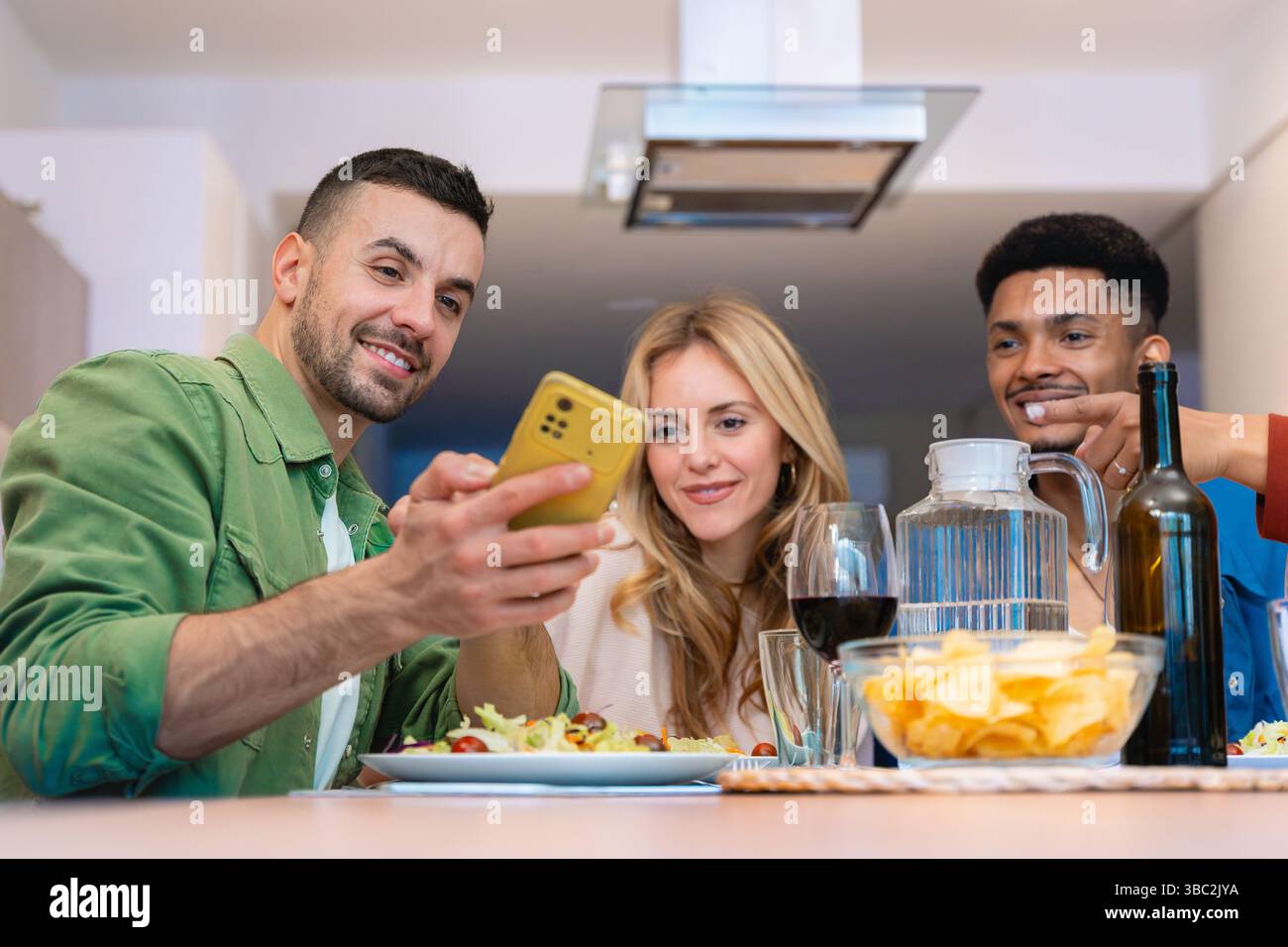Fröhliche multiethnische Freunde genießen Snacks und Getränke in moderner, heller Küche Stockfoto