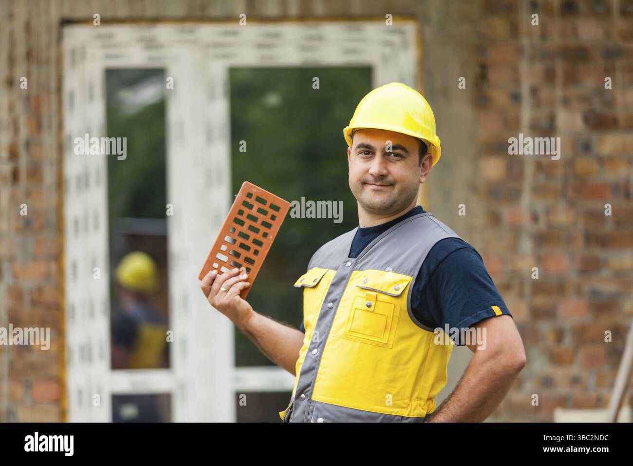 Der Mann mit gelber Weste und Helm steht auf dem Hintergrund eines unvollendeten Hauses und hält einen Ziegel in der Hand Stockfoto