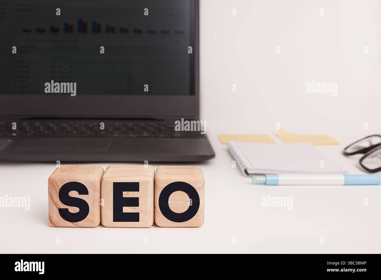 Holzblöcke mit der Schreibweise „SEO“ stehen im Mittelpunkt eines aufgeräumten Arbeitsplatzes. Ein Laptop, ein Notizblock, ein Stift und Haftnotizen sitzen im unscharfen Hintergrund. Stockfoto