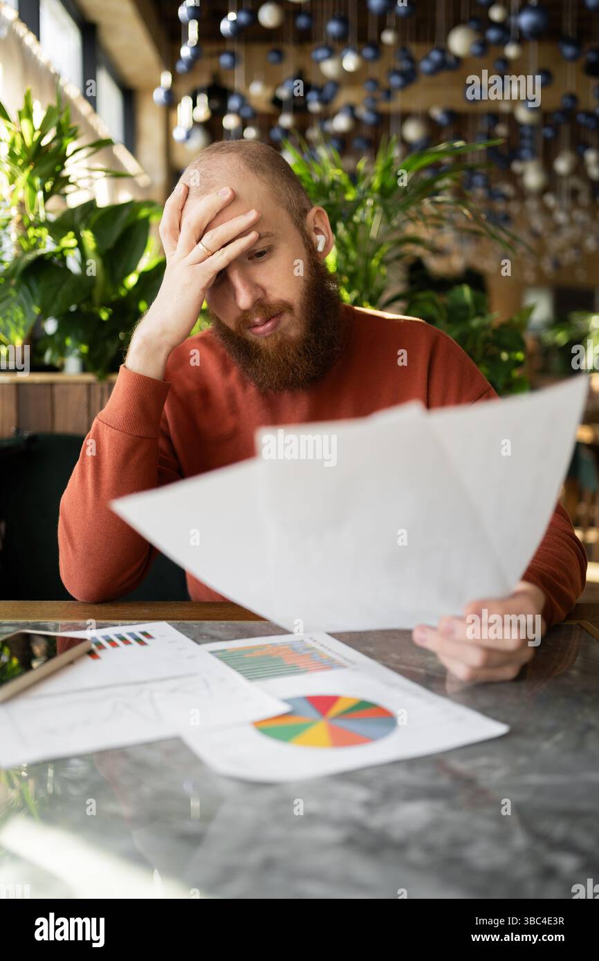 Junger bärtiger Buchhalter, der im Café Dokumente liest und den Kopf mit den Händen hält, Konzept von Problemen und Konkurs. Stockfoto
