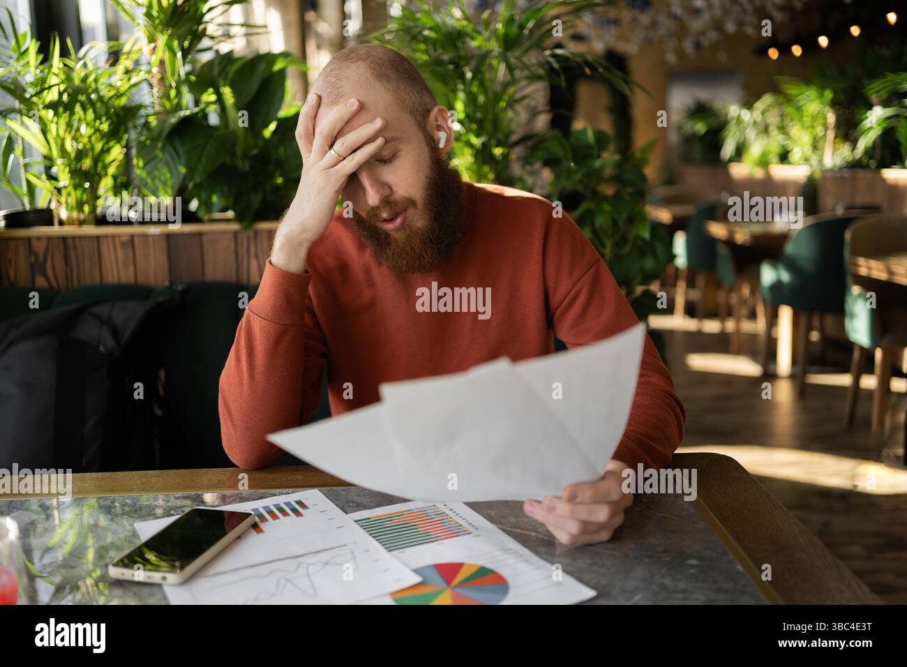 Junger bärtiger Buchhalter, der im Café Dokumente liest und den Kopf mit den Händen hält, Konzept von Problemen und Konkurs Stockfoto