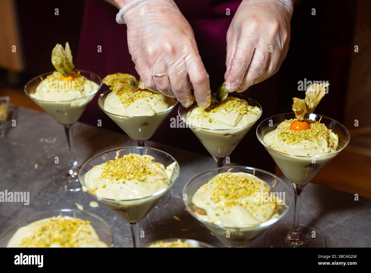 Mit Handschuhen dekorierte cremige Pistaziendesserts in Martini-Gläsern mit zerquetschten Nüssen und Physalis auf einer Küchentheke. Stockfoto