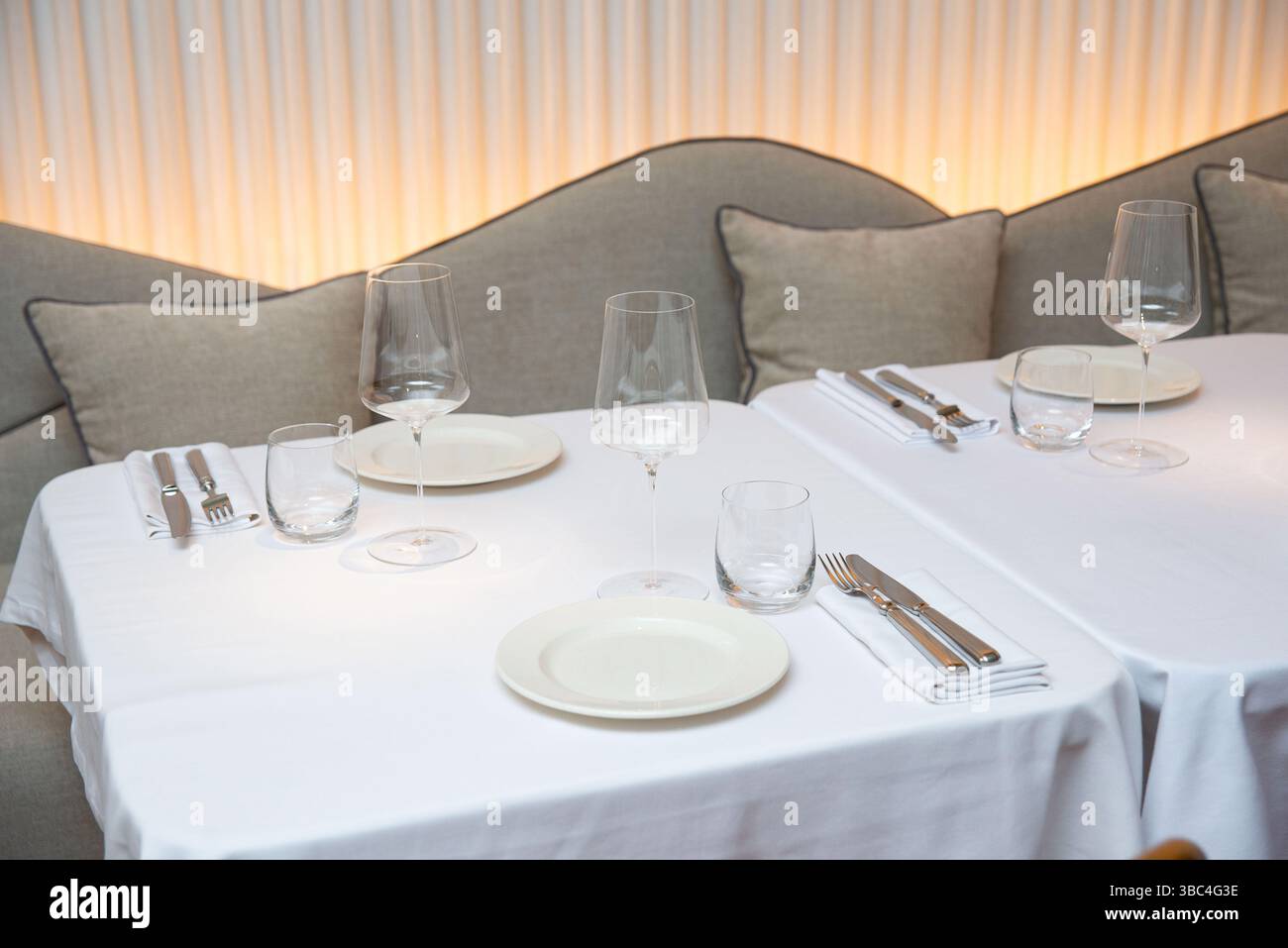 Ein ordentlich angeordneter Esstisch in einem modernen Restaurant mit weißer Bettwäsche, Weingläsern, Tellern und Besteck in gedämpfter Beleuchtung. Stockfoto