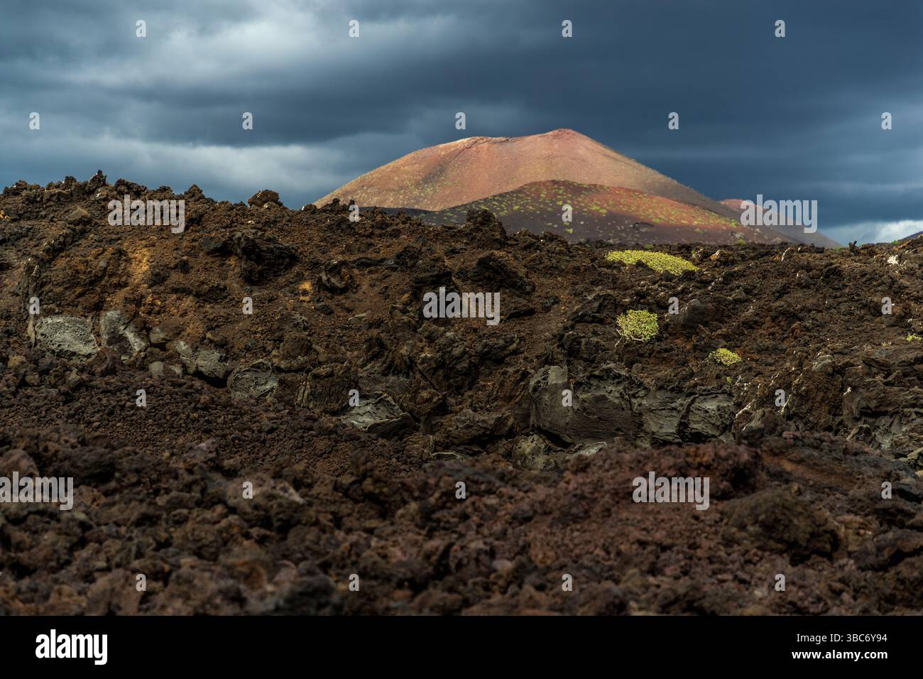 Die Montaña Bermeja erhebt sich rötlich über verfestigtem Lavagestein entlang der Avenida Marítima nahe El Golfo auf Lanzarote, Kanarische Inseln, Spanien. Ein einziger Ausbruch bildete den Kegel – auf Lanzarote bezeichnet „montaña“ einen Vulkan, der durch nur einen Ausbruch entstanden ist. Eisenhaltiger Vulkanboden verleiht ihm seine rostrote Farbe (bermejo), ein Merkmal, das mehreren Kegeln der Insel ihren Namen gibt. Gelb-grüner Flechtenbewuchs besiedelt das schwarze Lavafeld im Vordergrund. Dunkle Wolken ziehen über den Himmel. Stockfoto