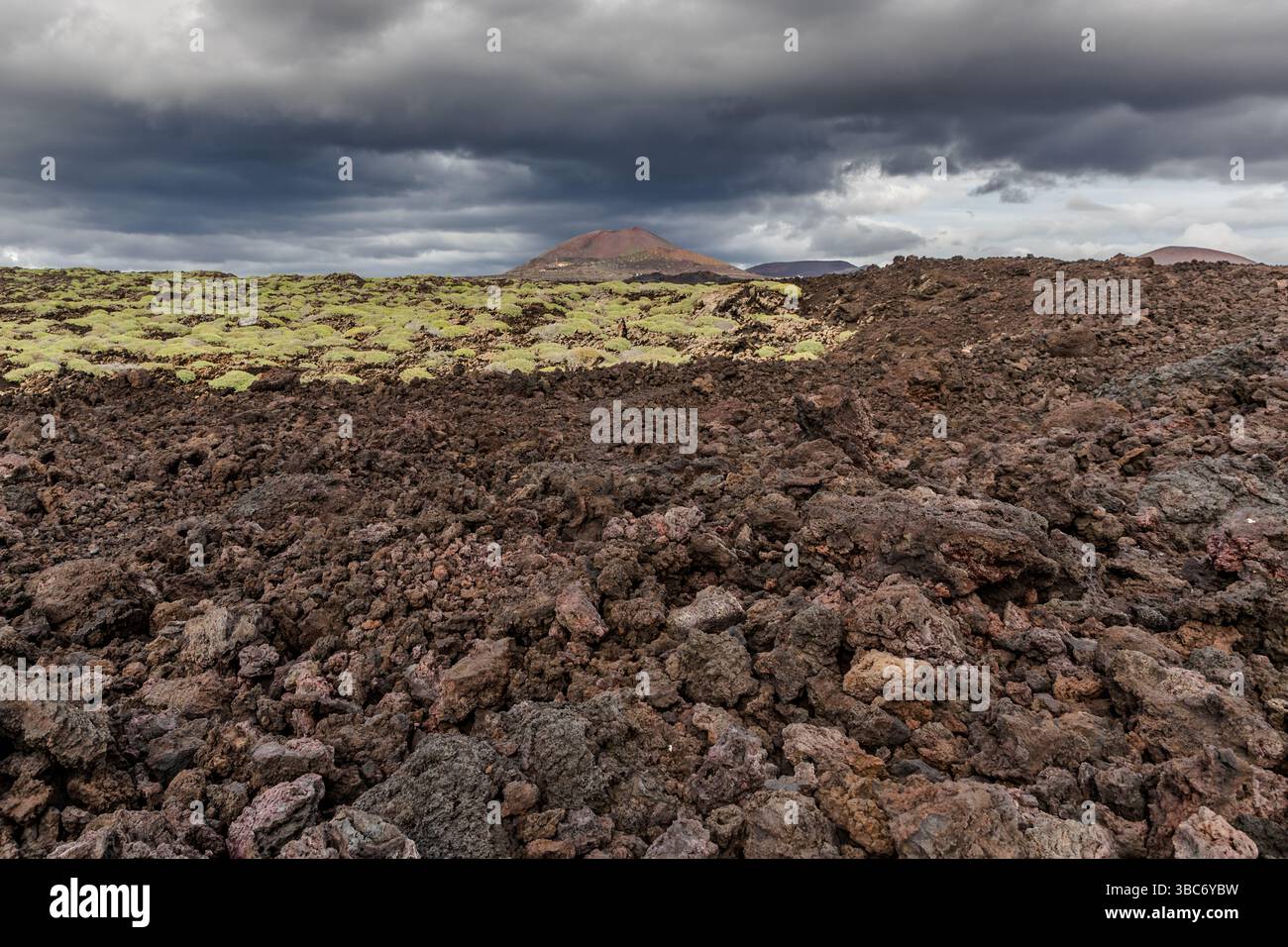 Die Montaña Bermeja erhebt sich rötlich im Hintergrund eines Lavafeldes entlang der Avenida Marítima in der Nähe von El Golfo auf Lanzarote, Kanarische Inseln, Spanien. Eine einzige Eruption bildete den Kegel – auf Lanzarote bezeichnet „montaña“ einen Vulkan, der durch nur eine Eruption entstanden ist. Eisenerreicher vulkanischer Boden verleiht ihm seine rostrote Farbe (bermejo), ein Merkmal, das mehreren Kegeln der Insel ihren Namen gibt. Gelbgrüner Flechten bedecken einen Streifen des schwarzen Lavafeldes. Dunkle Wolken hängen über der Szene. Stockfoto