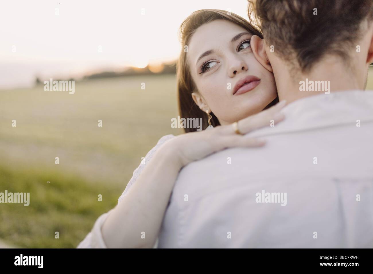 Eine Frau schaut, während sie einen Mann auf einem Feld umarmt, während sie bei Sonnenuntergang Romantik und Gelassenheit vermittelt Stockfoto