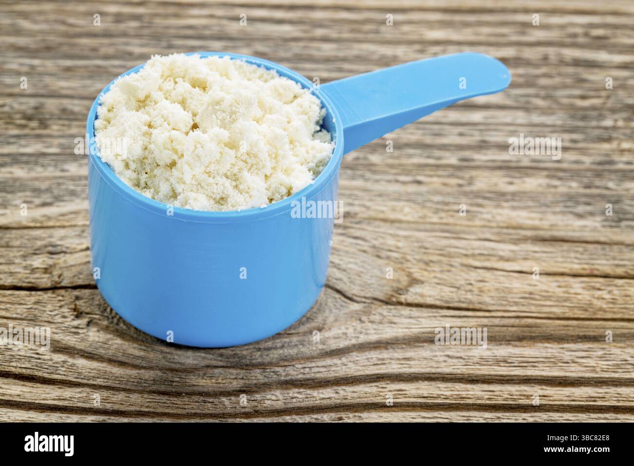 Molkenproteinpulver in einem blauen Kunststoff-Messlöffel gegen gemasertes Holz Stockfoto