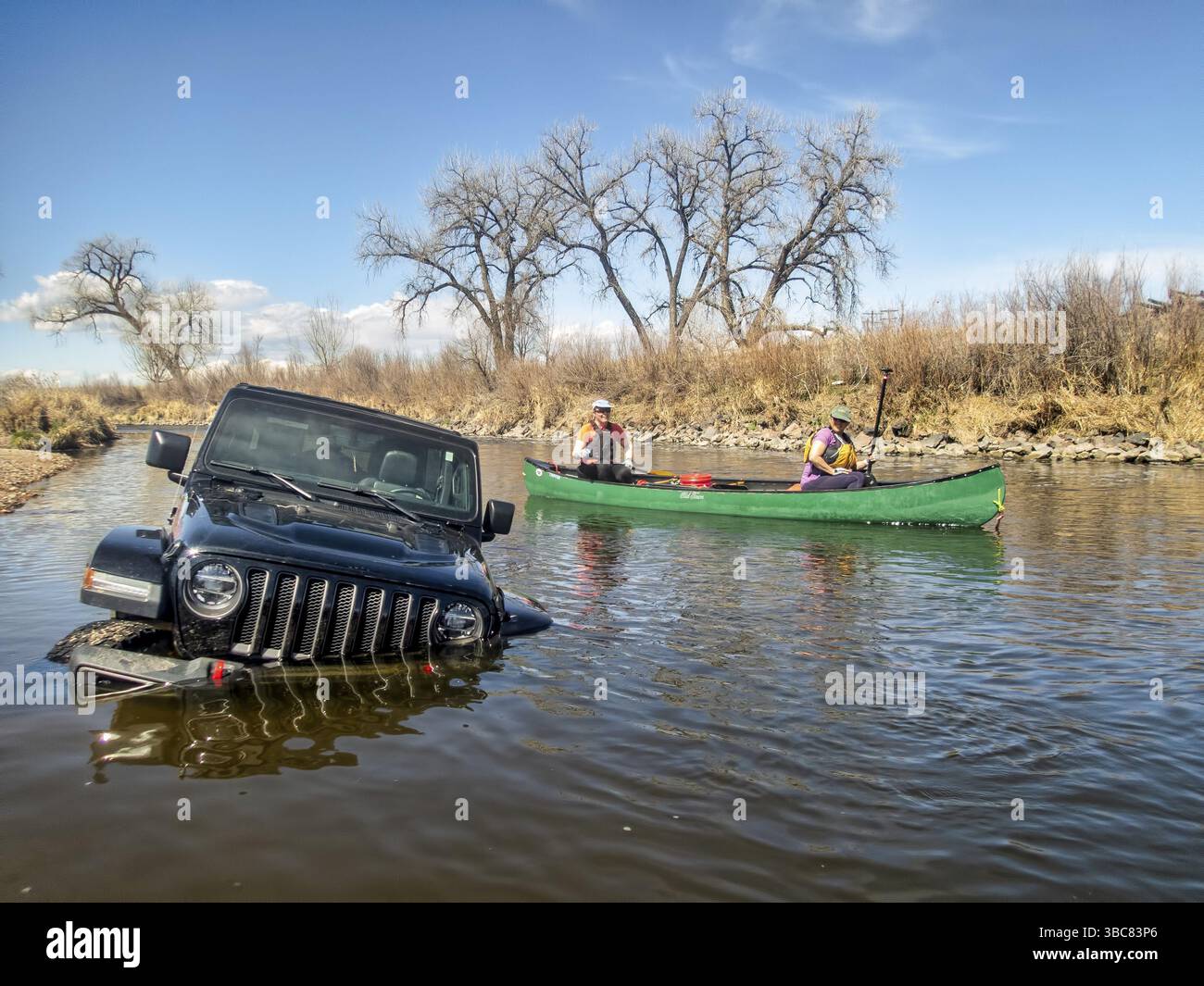 Brighton, CO, USA - 6. April 2019: Frauen in einem Kanu fahren am Jeep Wrangler vorbei, der tief im Fluss steckt, und die Frühjahrslandschaft am South Platte River Stockfoto