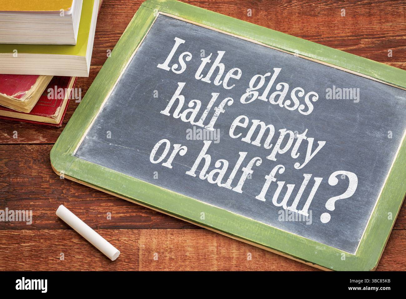 Ist das Glas halb leer oder halb voll Frage auf einer Schiefertafel mit einer weißen Kreide und einem Stapel Bücher vor einem rustikalen Holztisch Stockfoto