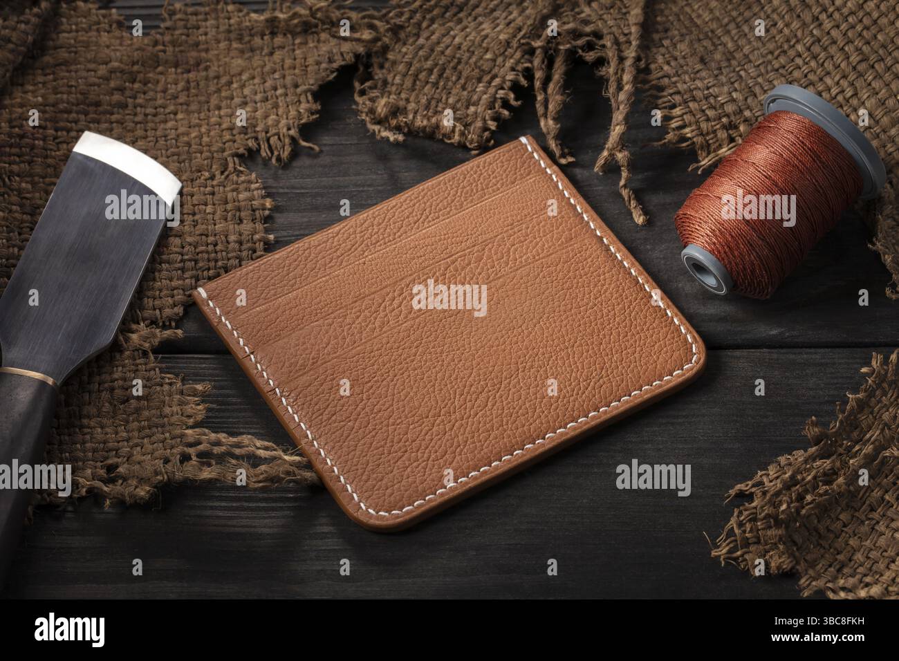 Ledertaschen herstellen. Eine Ledertasche, eine Spule mit Faden und ein Messer liegen auf einem Holztisch Stockfoto