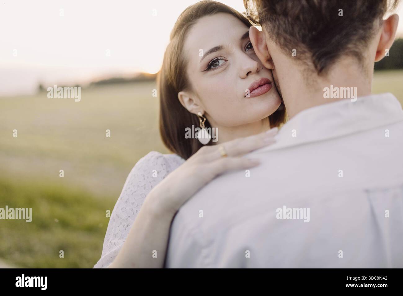 Nahaufnahme einer Frau, die über die Schulter ihres Partners schaut, während die Sonne auf einem Feld untergeht und einen zarten Moment festnimmt Stockfoto