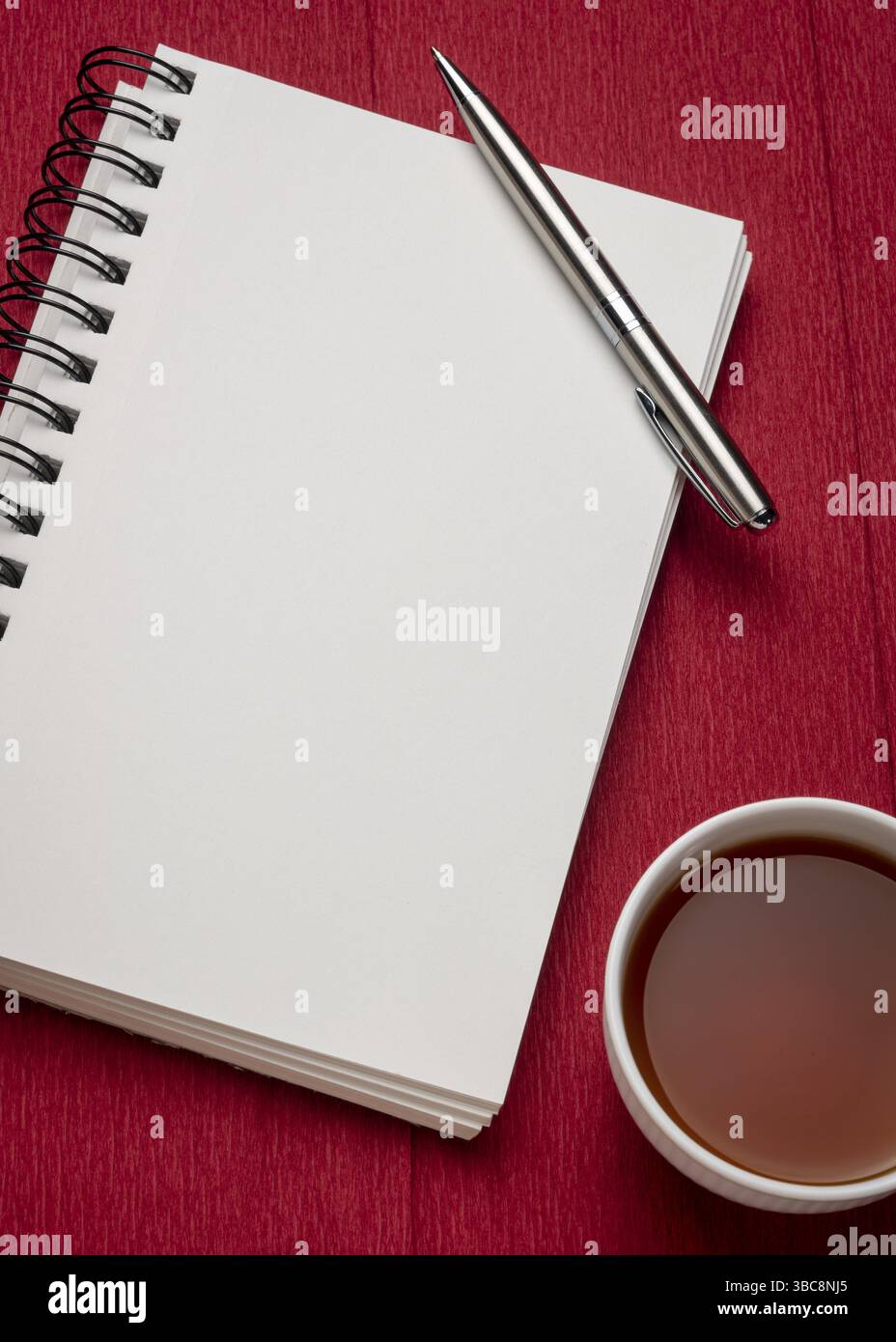 Leeres spiralförmiges Kunstskizzenbuch mit einer Tasse Tee gegen strukturiertes rotes Krepppapier, Arbeitsplatzkonzept Stockfoto