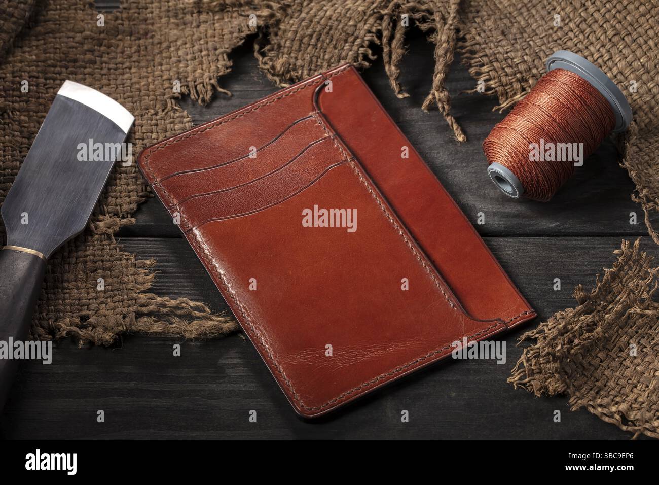 Ledertaschen herstellen. Eine Ledertasche, eine Spule mit Faden und ein Messer liegen auf einem Holztisch Stockfoto