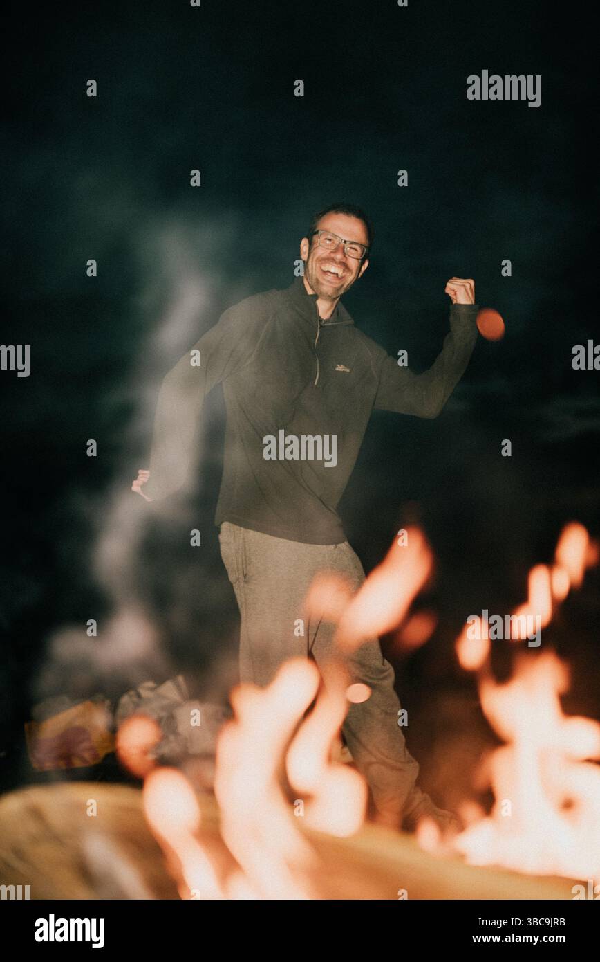 Ein Mann steht vor einem Feuer, lächelt und winkt. Das Feuer ist groß und hell und schafft eine warme und einladende Atmosphäre. Der Ausdruck des Mannes Stockfoto