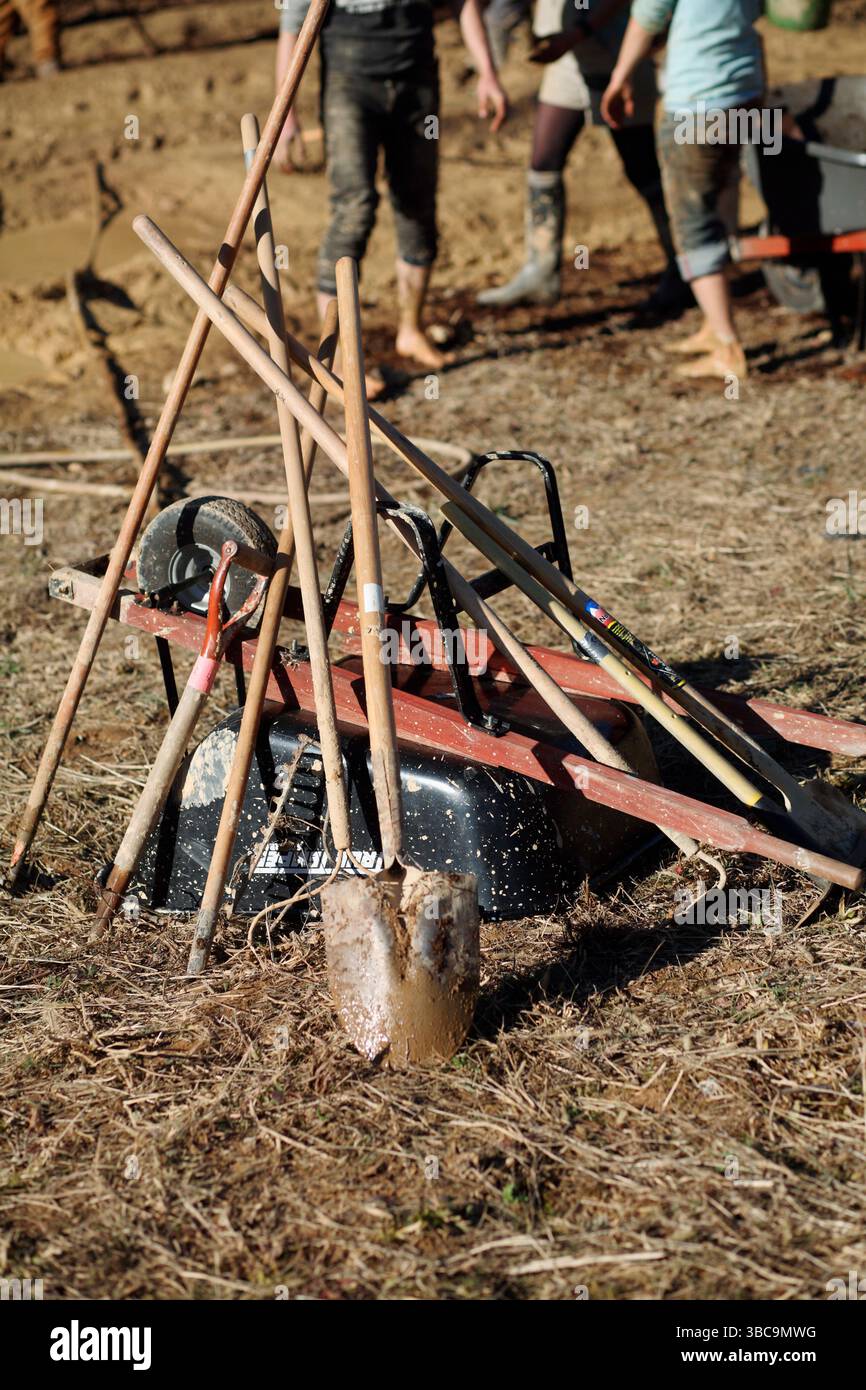 Grabwerkzeuge – Schaufeln, Meißelachsen und Schubkarren – gestapelt bei einem Erntegutmob Stockfoto