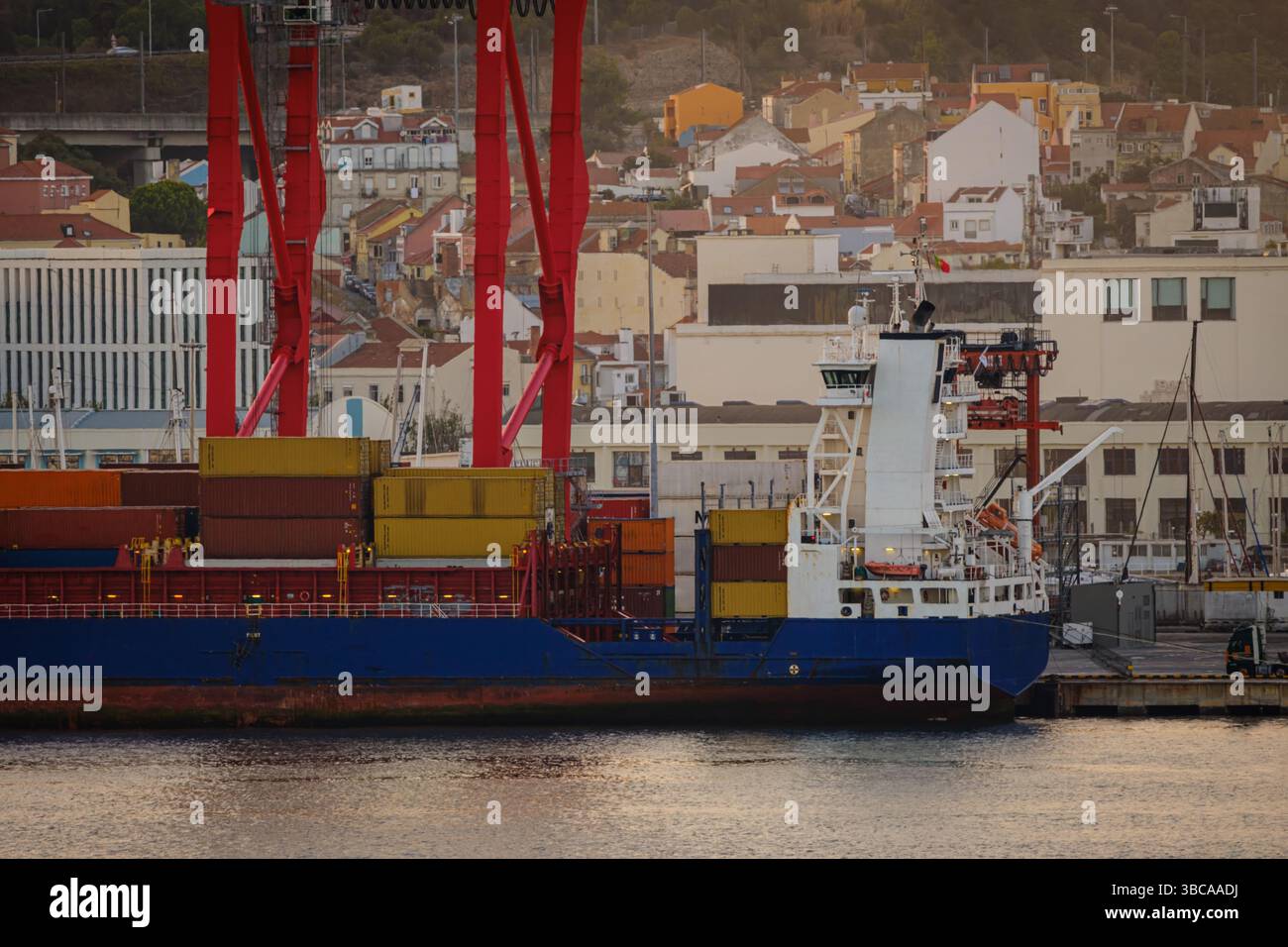Frachtschiff beladen mit bunten Containern im Hafen von Lissabon, roten Kränen und pulsierenden Gebäuden der Stadt am sonnigen Abend. Dynamische Wohn- und Industrieszene durch Uferpromenade, städtische Skyline, Be- und Entladen, ruhiges Wasser, Handel, Industrie Stockfoto