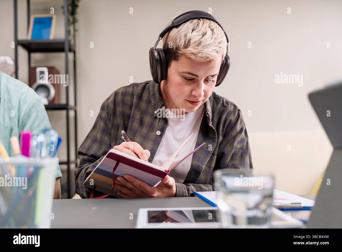 Lässiger Millennials-Profi, der Kopfhörer trägt und Ideen in einem gestreiften Notebook an einem modernen Coworking-Schreibtisch schreibt, Konzept von Fokus, Studium und Crea Stockfoto