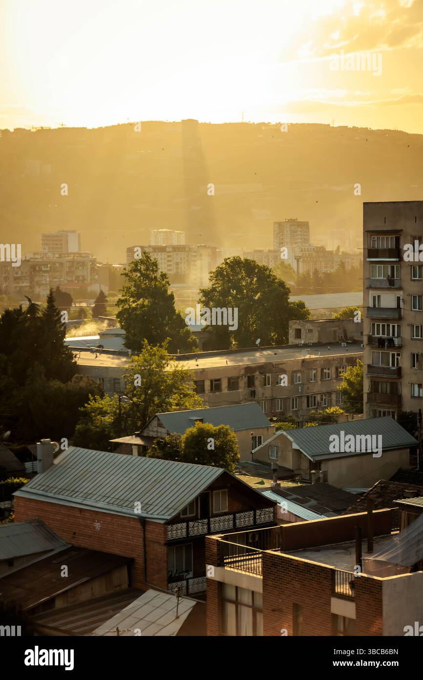 Goldenes Sonnenlicht strömt durch Wolken über die Stadtlandschaft von Tiflis und wirft bei Sonnenuntergang dramatische Lichtstrahlen über Wohngebäude und Bäume. Warm, bei Stockfoto