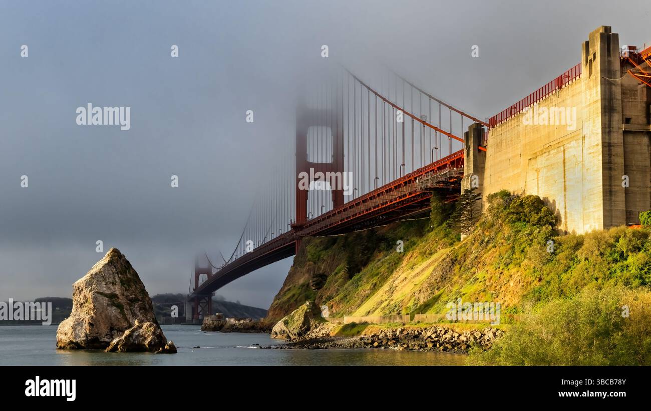 Eine majestätische rote Hängebrücke erhebt sich aus dem Nebel, deren hohe Türme teilweise verdeckt sind. Sonnenlicht beleuchtet die felsige Küste und grüne hil Stockfoto