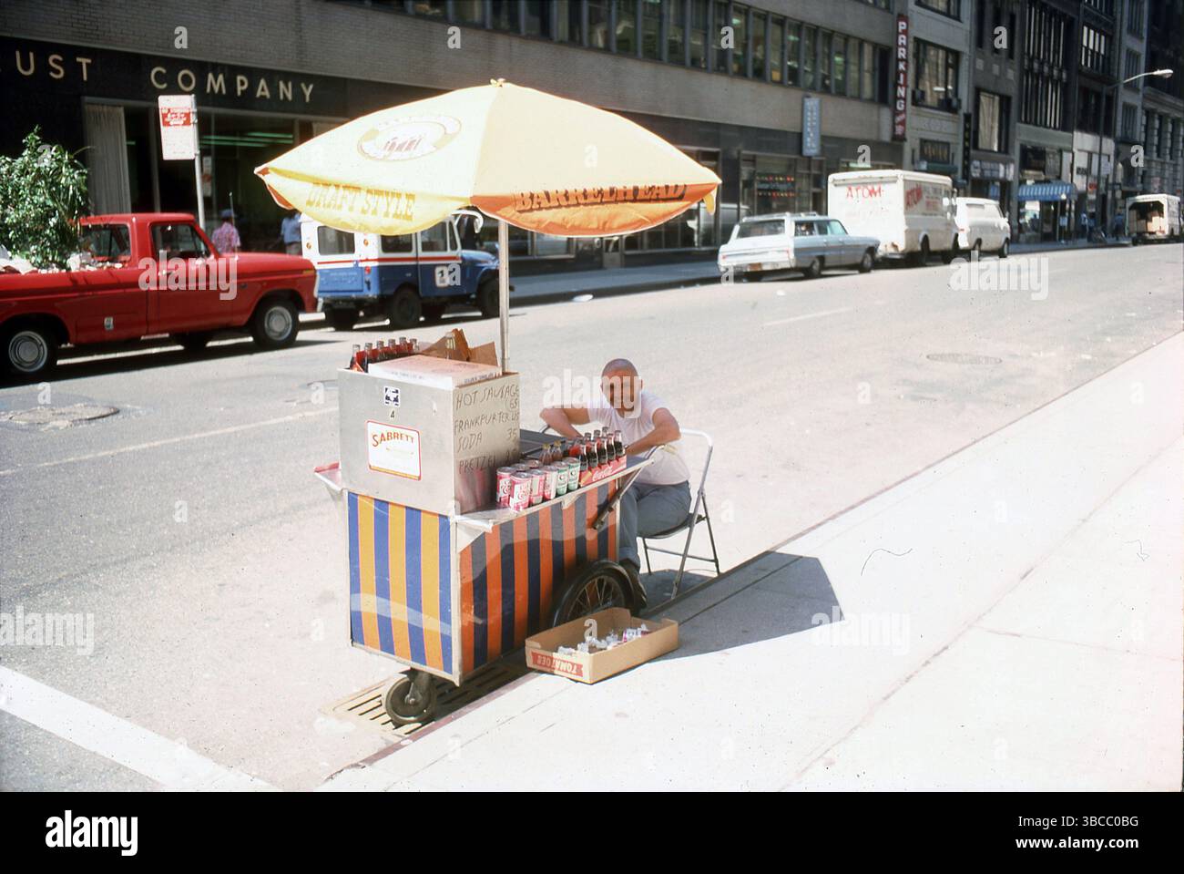 1978, Sommer in New York City und am Straßenrand, ein Hot Dog Verkäufer sitzt an seinem mobilen Stand in New York, NY, USA. Auf dem handgeschriebenen Menü steht: Warme Wurst 65 Cent, Frankfurter 45 Cent, Soda 35, Brezel 25 Cent Stockfoto