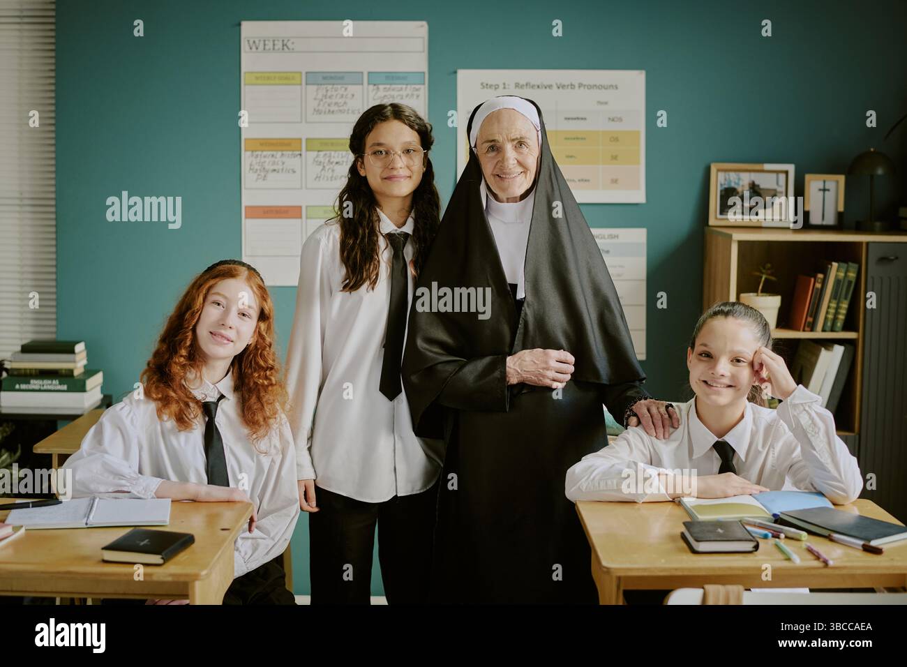 Porträt einer lächelnden Nonne, die mit drei Schulkindern an Schreibtischen im Klassenzimmer steht, mit pädagogischen Plakaten und Bücherregalen im Hintergrund Stockfoto