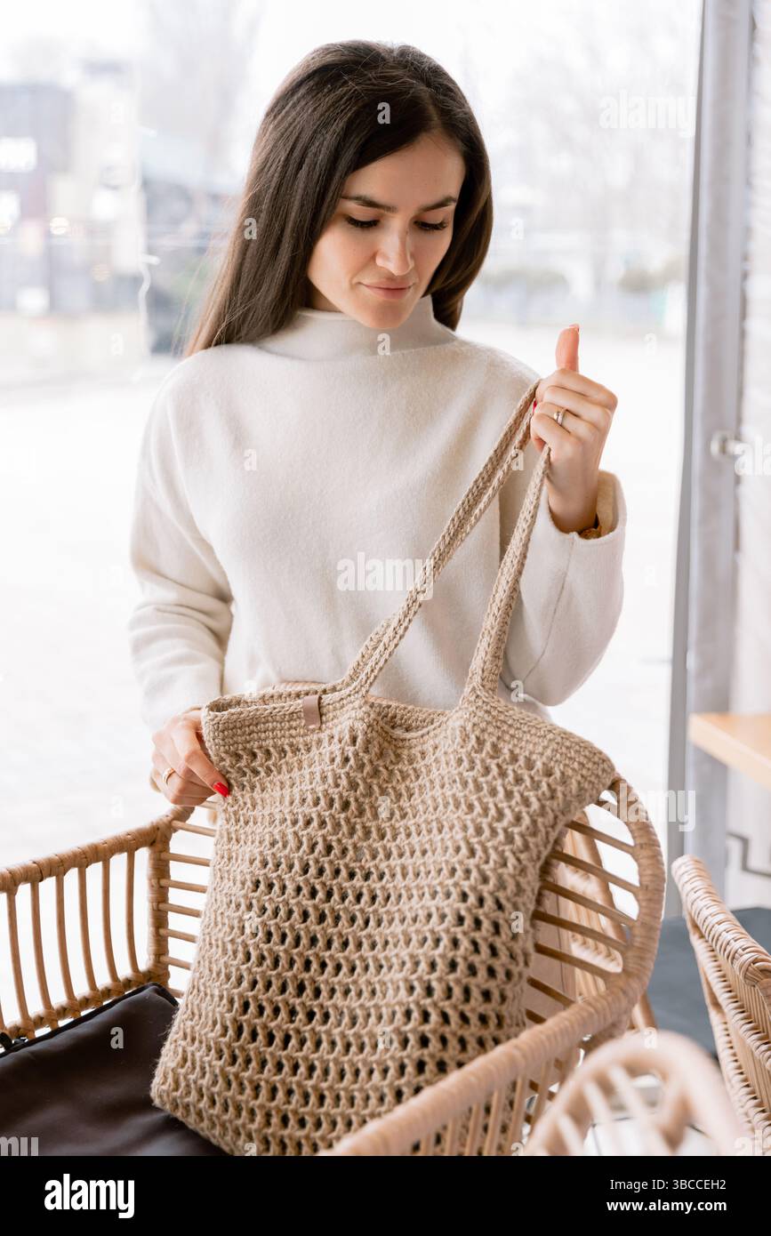 Stilvolle Frau mit roten Nägeln, die eine umweltfreundliche gewebte Tasche in einem gemütlichen, modernen Café mit natürlichem Dekor hält. Stockfoto