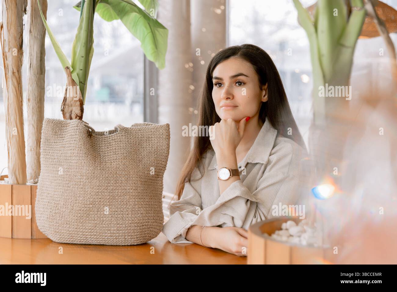 Stilvolle Frau, die sich in einem gemütlichen Café mit natürlichem Dekor entspannt, mit eleganter Uhr und Leinen-Outfit. Stockfoto