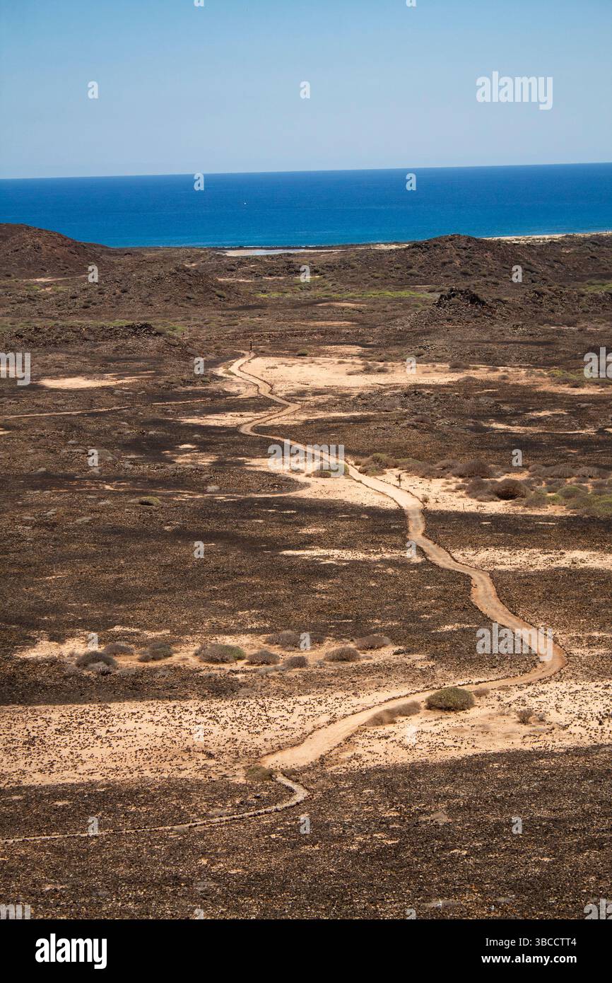 Ein kurviger Pfad führt durch das trockene vulkanische Gelände und erstreckt sich zum blauen Ozean am Horizont. Die mäandernde Form der Straße repräsentiert das t des Lebens Stockfoto