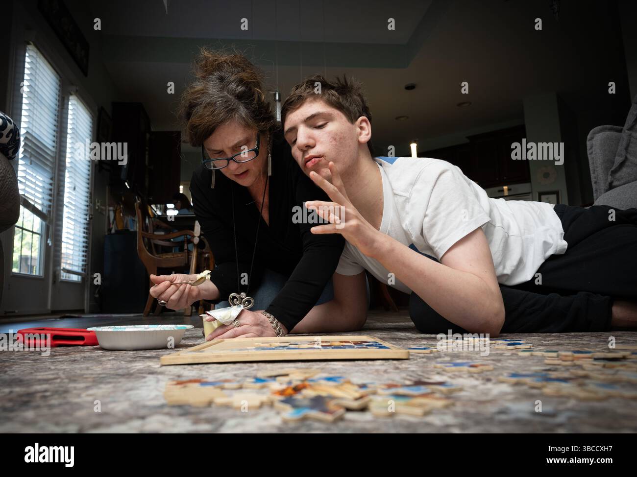Ft. Mill, South Carolina, USA. März 2025. Beth Hodges schließt sich ihrem 19-jährigen Sohn Jack auf dem Wohnzimmerboden an, während er mit einem einfachen Puzzle spielt und ihm einen Puddingsnack füttert. Er hat einen seltenen Krampfanfall namens Dravet-Syndrom sowie Autismus der Stufe 3. (Kreditbild: © Robin Rayne/ZUMA Press Wire) NUR REDAKTIONELLE VERWENDUNG! Nicht für kommerzielle ZWECKE! Stockfoto