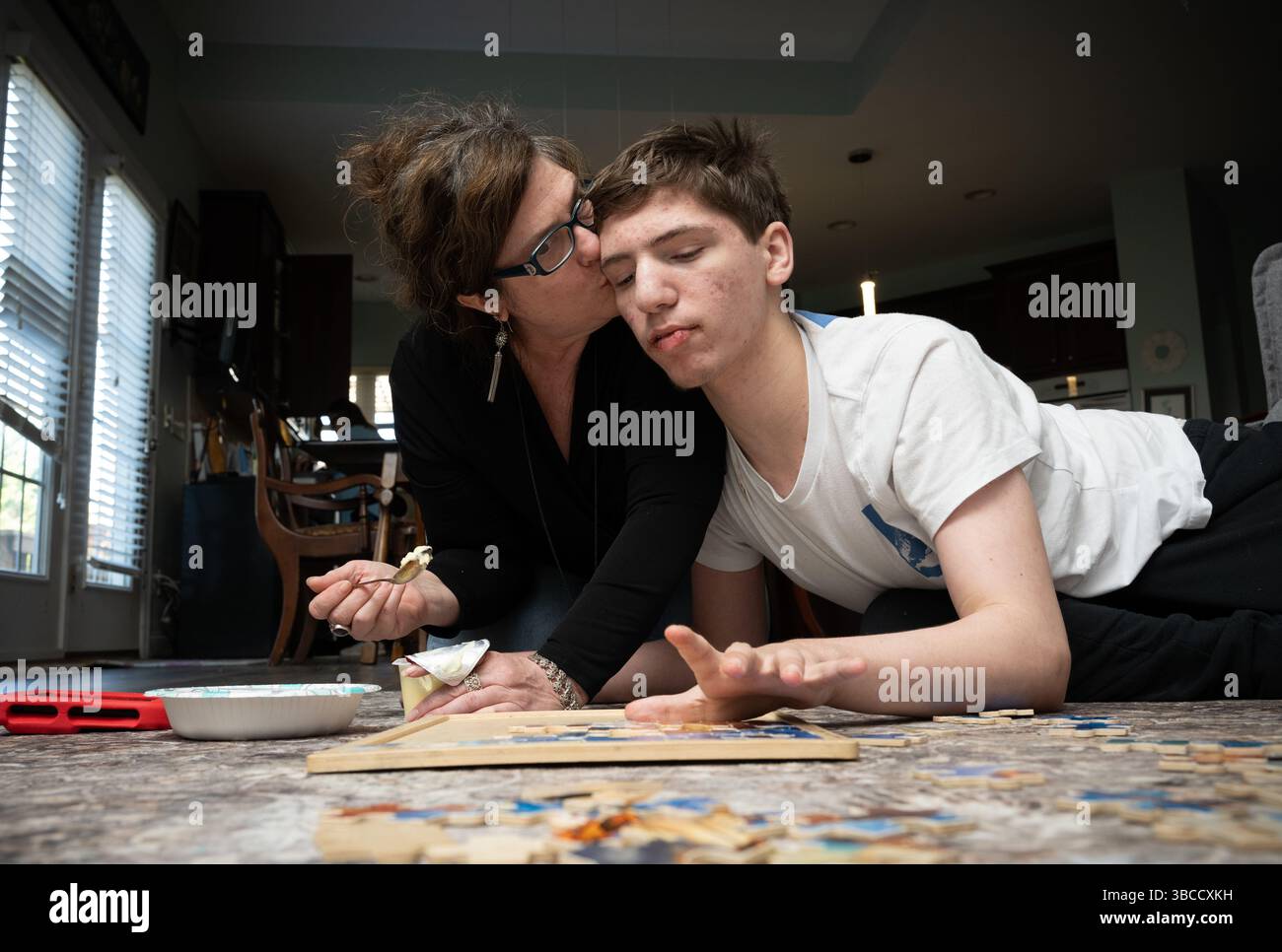 Ft. Mill, South Carolina, USA. März 2025. Beth Hodges schließt sich ihrem 19-jährigen Sohn Jack auf dem Wohnzimmerboden an, während er mit einem einfachen Puzzle spielt und ihm einen Puddingsnack füttert. Er hat einen seltenen Krampfanfall namens Dravet-Syndrom sowie Autismus der Stufe 3. (Kreditbild: © Robin Rayne/ZUMA Press Wire) NUR REDAKTIONELLE VERWENDUNG! Nicht für kommerzielle ZWECKE! Stockfoto
