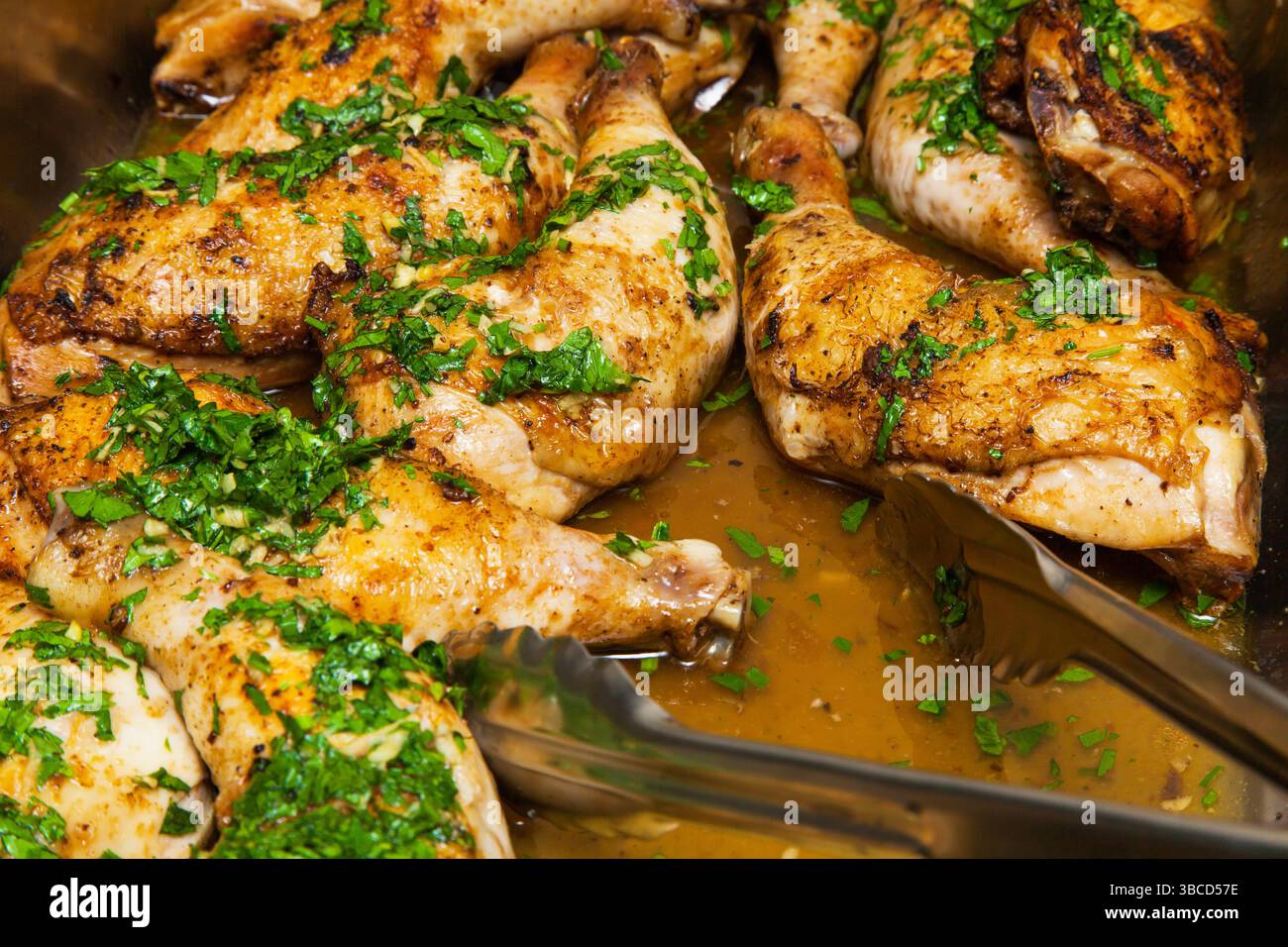 Gebackene Hähnchenschenkel in eigenem Fett, bestreut mit gehackter Petersilie und Knoblauch in einer Schüssel mit einer Zange Nahaufnahme. Stockfoto