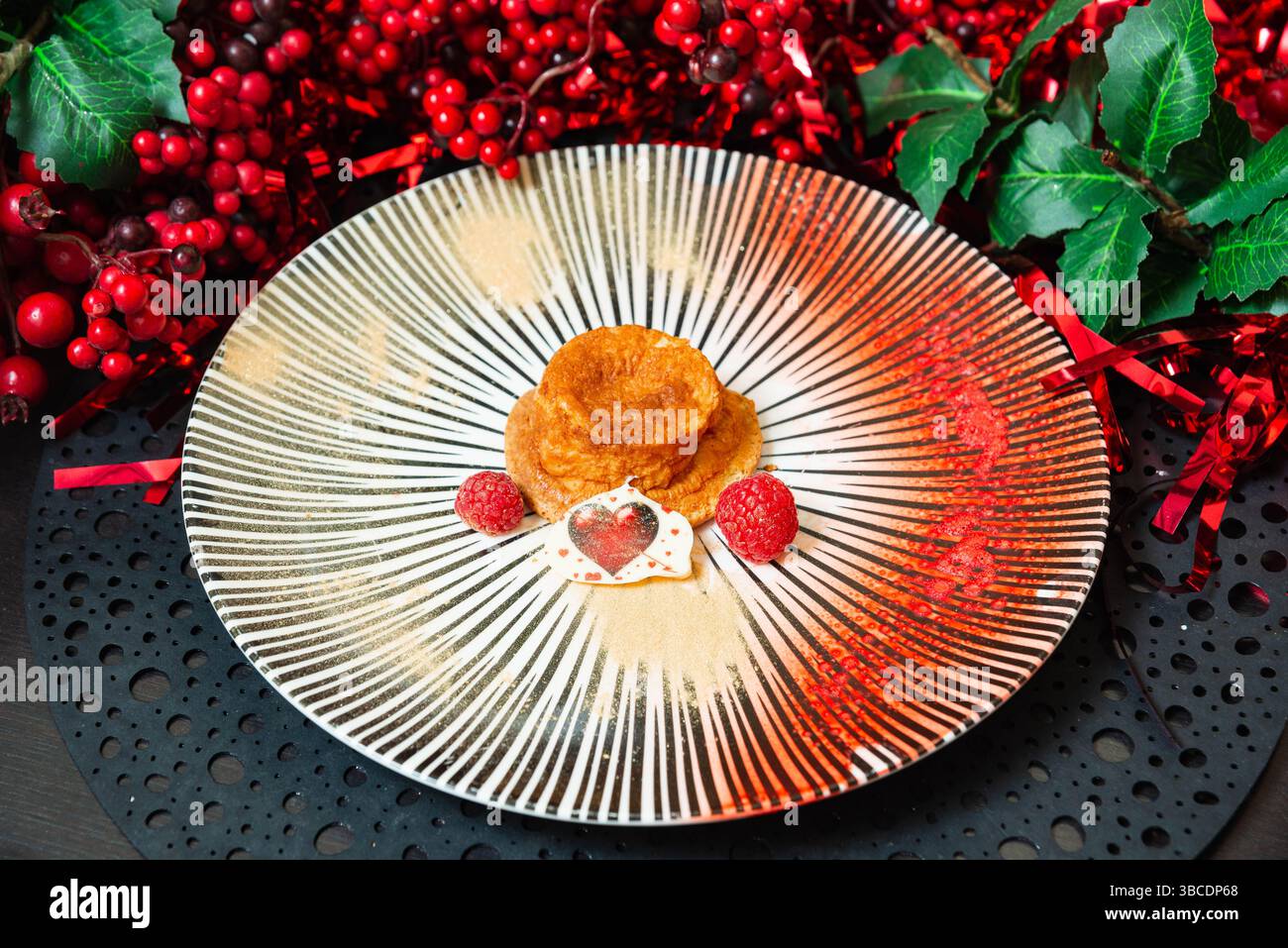 Festliches Dessert mit einem kleinen Kuchen, frischen Himbeeren und herzförmiger Schokolade, dekoriert mit rotem Dekor für eine Weihnachtsfeier. Stockfoto