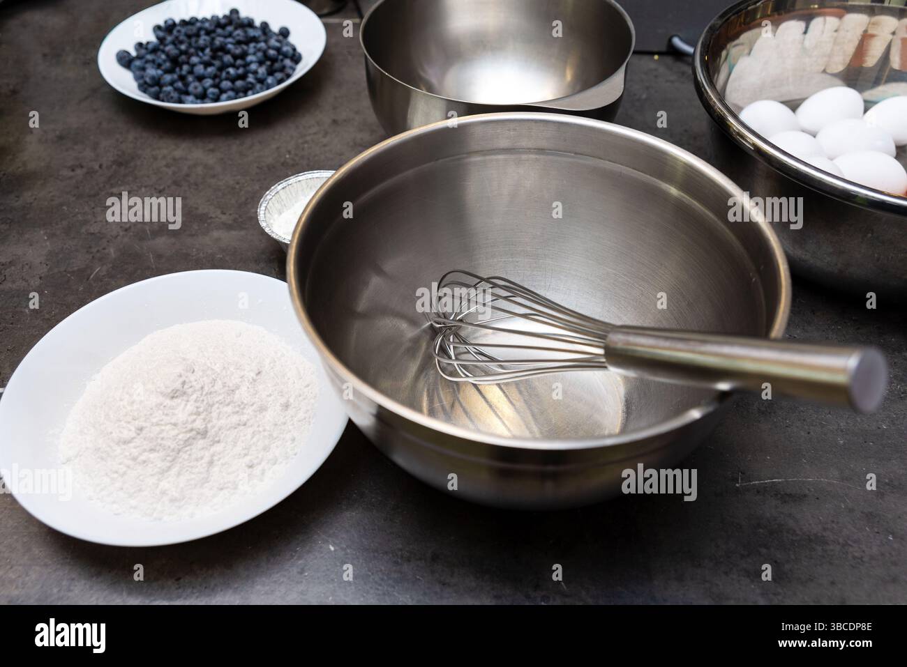 Rührschüssel aus Edelstahl mit Schneebesen, umgeben von Backzutaten, einschließlich Mehl, Eiern und frischen Heidelbeeren, bereit für die Rezeptzubereitung. Stockfoto