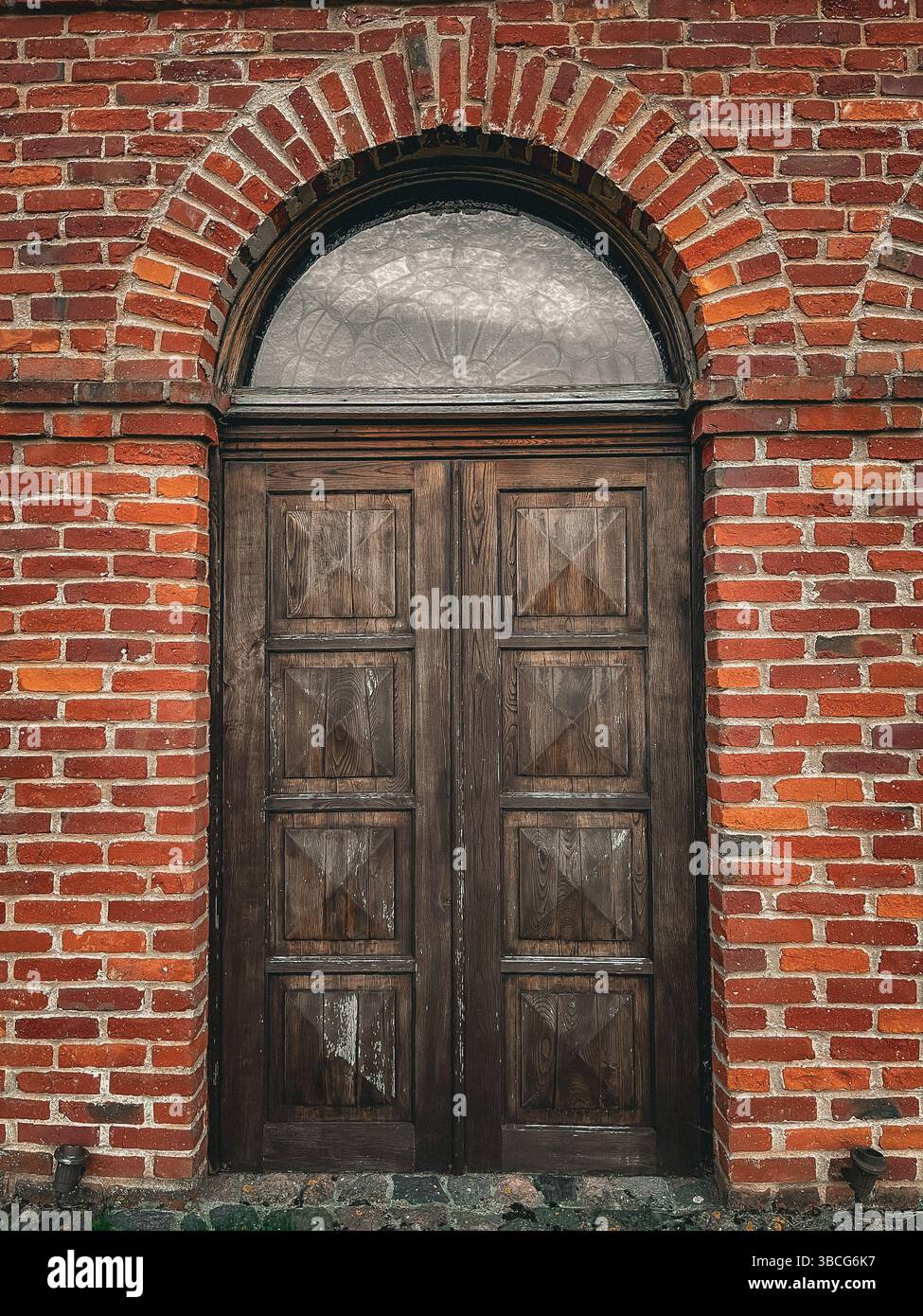 Eine alte Holzdoppeltür in einer roten Ziegelwand mit einem gewölbten Milchfenster darüber, die rustikale europäische Architekturdetails zeigt. Stockfoto