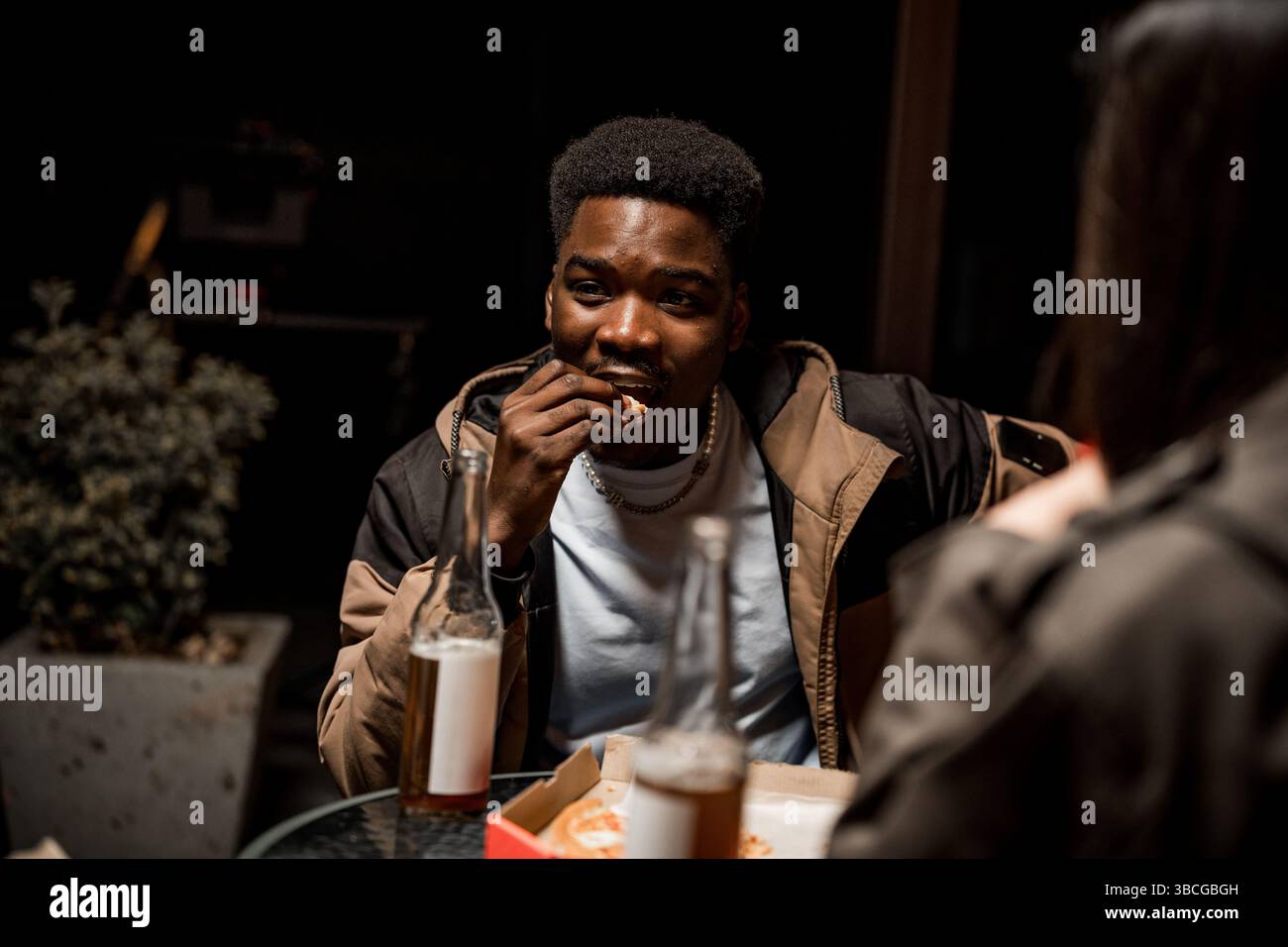 Junger Mann, der abends mit Freunden im Freien einen Snack genießt, während er Drinks trinkt und sich unterhält Stockfoto