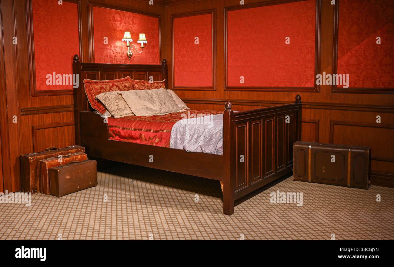Elegantes Vintage-Schlafzimmer mit Holzbett, Koffern und roter Wandverkleidung, die an ein luxuriöses Reiseerlebnis erinnern. Stockfoto
