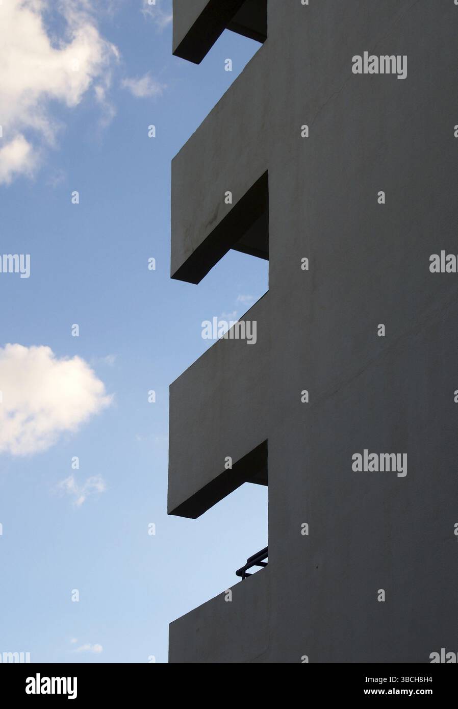 Die Ecke eines hohen Apartmentgebäudes mit Balkonen, die geometrische Formen vor einem blauen, bewölkten Himmel bilden Stockfoto
