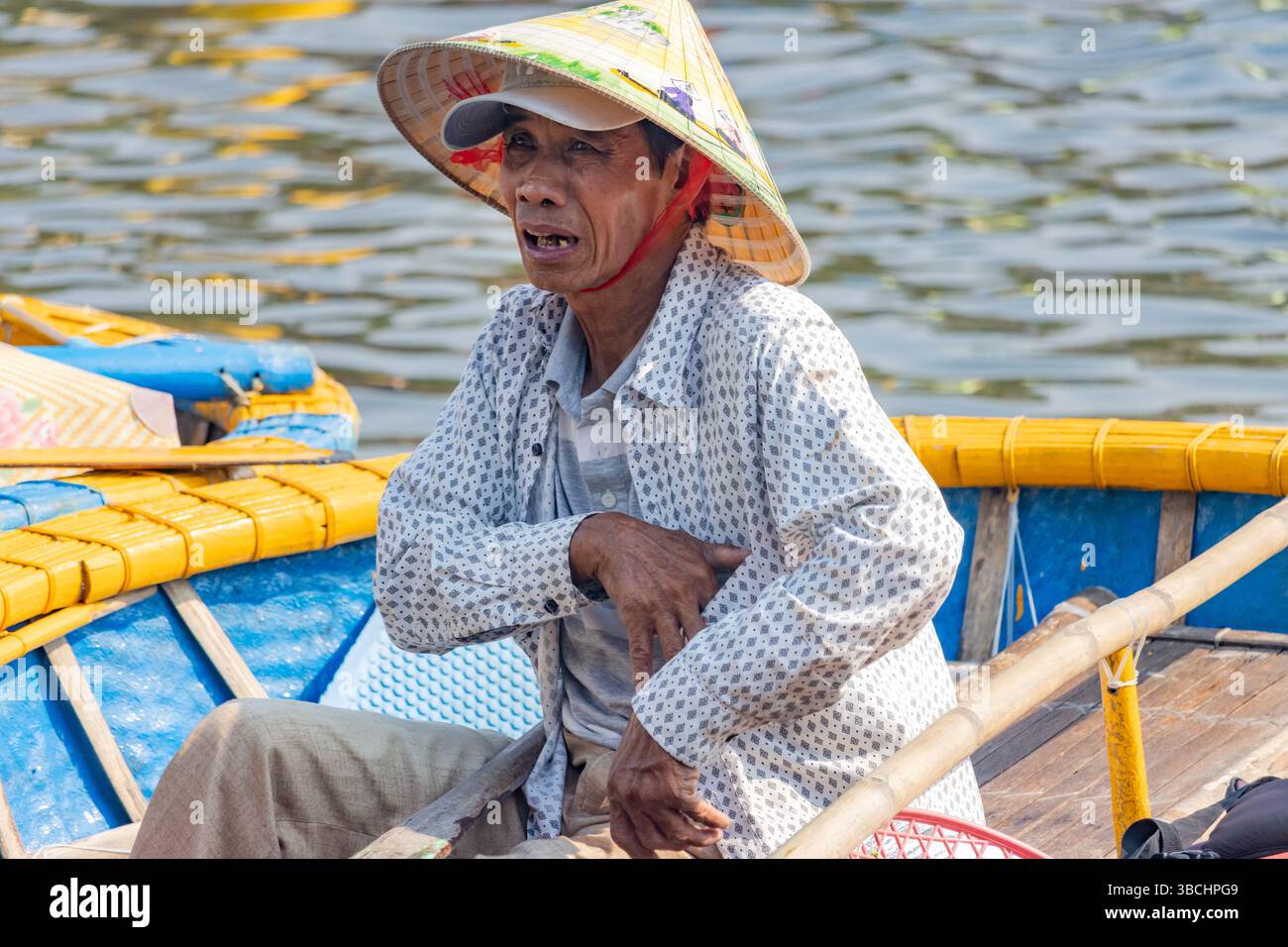 HOI AN, VIETNAM, 26. April 2025, Ein Mann mit traditionellem Strohhut und gemustertem Hemd, der auf einem bunten Holzboot sitzt Stockfoto