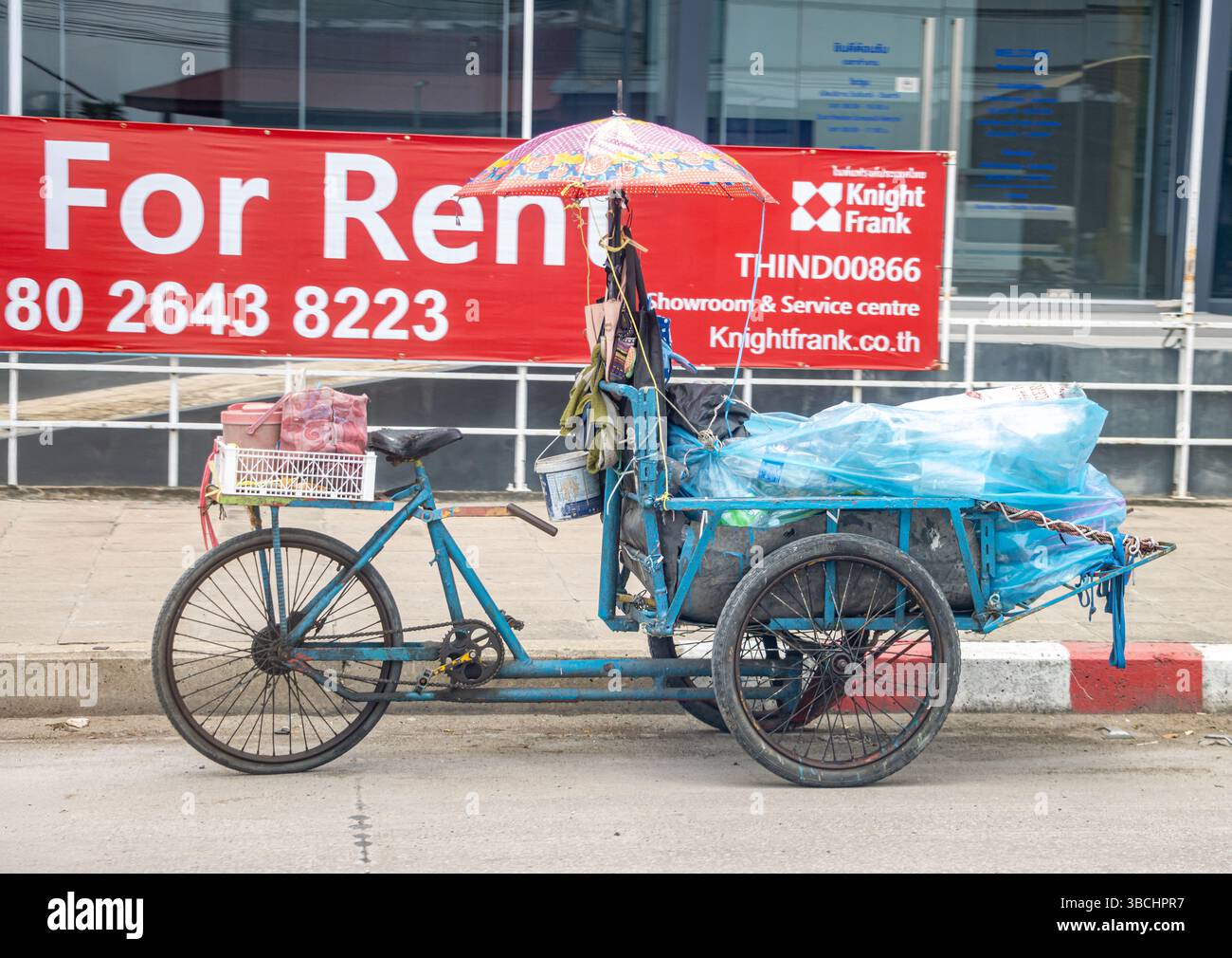 SAMUT PRAKAN, THAILAND, 04. MAI 2025, Vintage-Dreiradwagen mit Ladung und Regenschirm neben dem städtischen Bürgersteig unter einer Werbetafel mit Werbung A geparkt Stockfoto