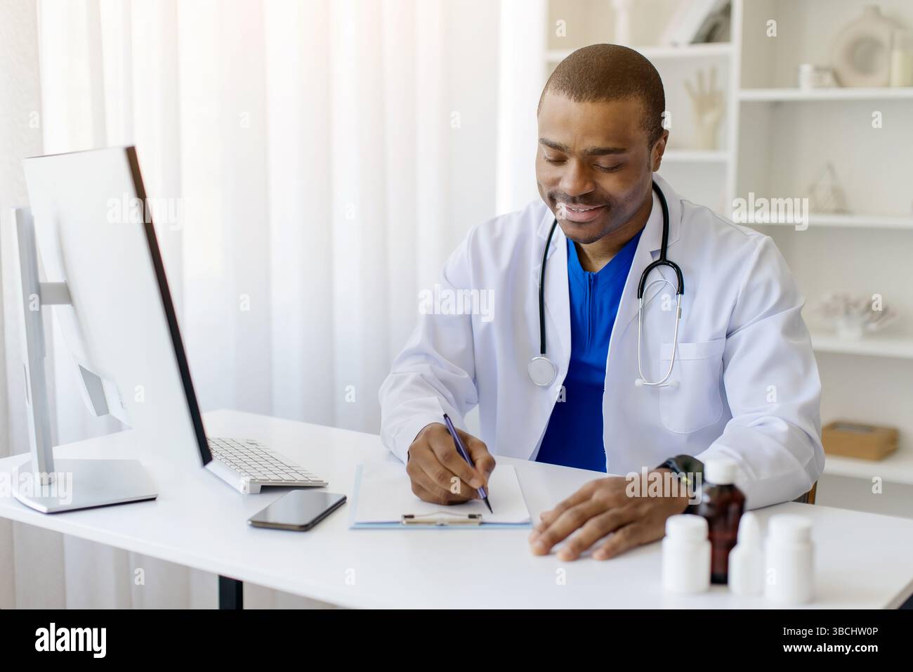 Glücklicher schwarzer Doktor, der am Schreibtisch in der Klinik sitzt und Notizen macht Stockfoto